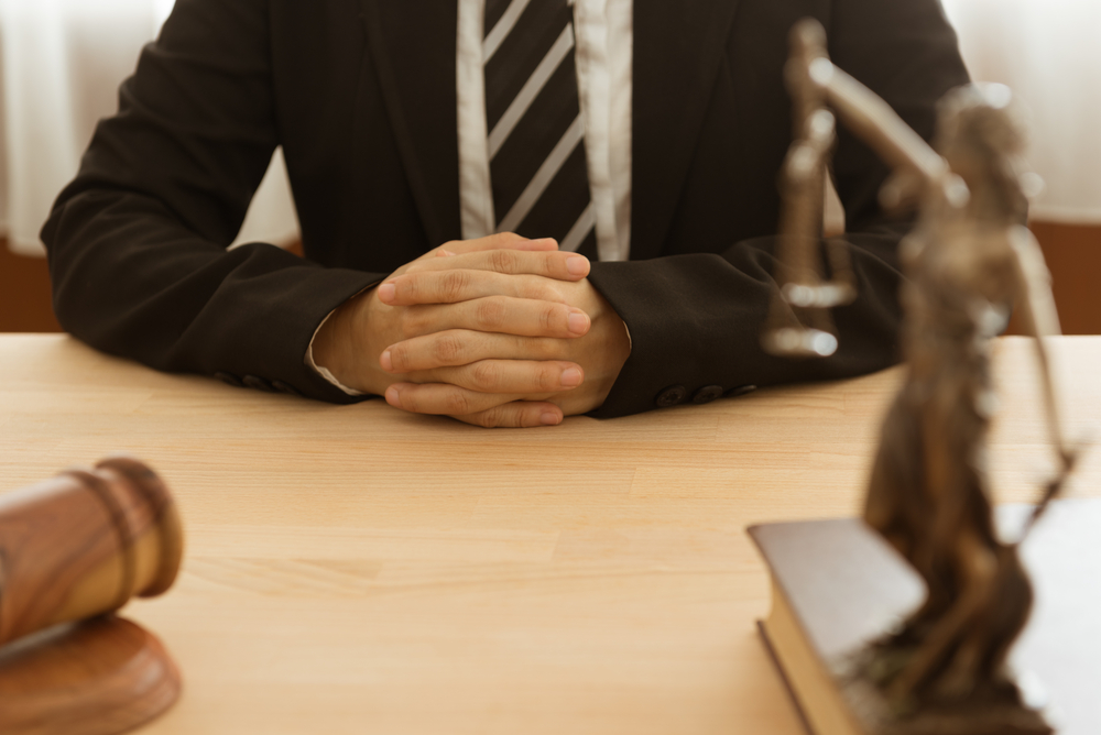 man in suit sits at table which holds judge's gavel and Lady Justice